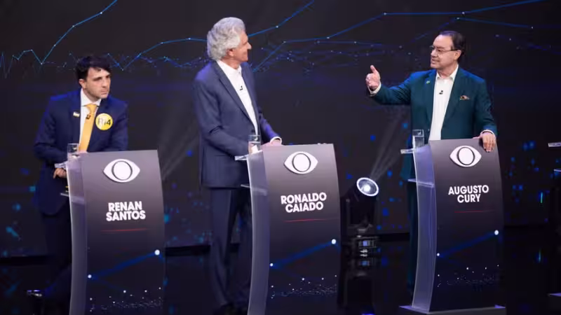 Morada FM — O primeiro debate presidencial das eleições de 2026 foi realizado no domingo (23), com participação de Ronaldo Caiado (PSD), Renan Santos (Missão) e Augusto Cury (Avante). Lula (PT) não compareceu e concedeu entrevista à Record no mesmo horário. Flávio Bolsonaro (PL) havia condicionado sua presença à participação do petista. Romeu Zema (Novo) também desistiu, alegando que a mudança de data perdeu o propósito com a ausência de Lula e de outros candidatos. Augusto Cury chegou a anunciar que não participaria, mas voltou atrás. O encontro foi considerado um dos mais esvaziados desde 1989. Renan Santos adotou tom agressivo e atacou os [&hellip;]